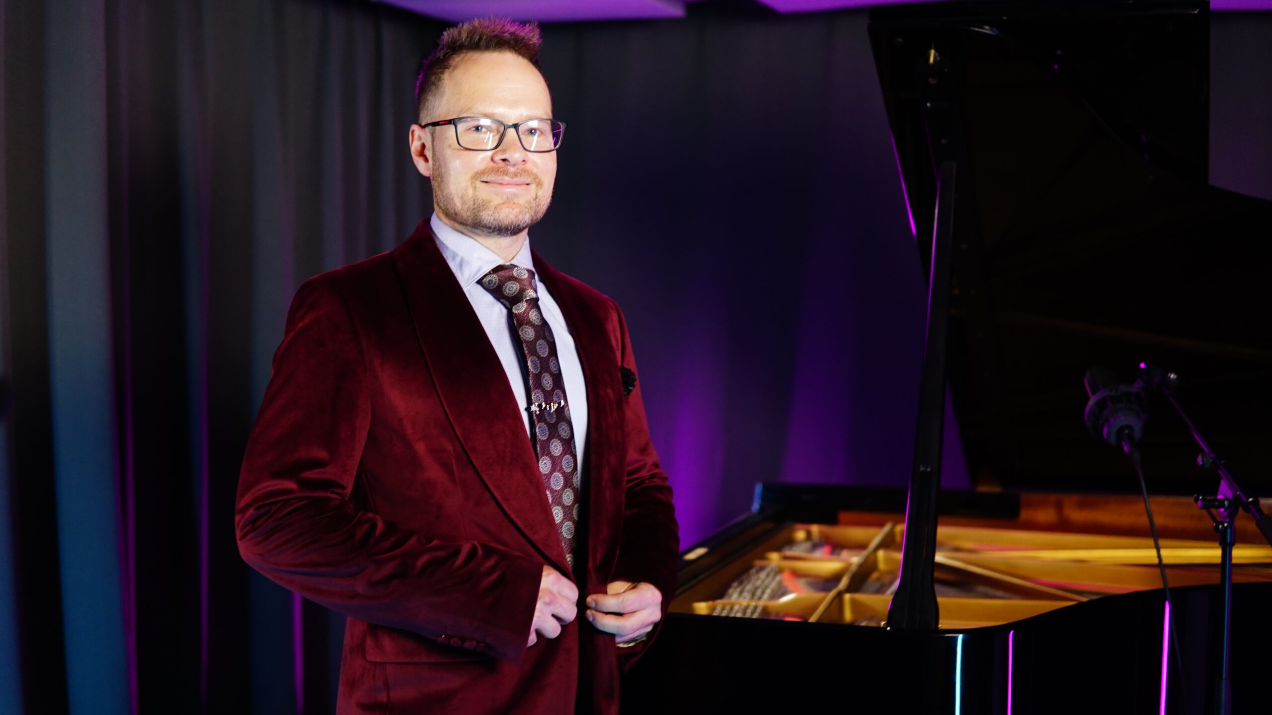 Simon Galfe in a burgundy velvet jacket beside an open black grand piano